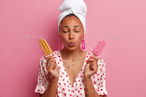 Isolated shot of satisfied young woman closes eyes and keeps lips rounded, poses with two ice creams