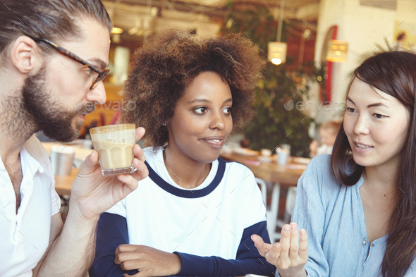 Three people of diverse races having conversation at cafe: Caucasian man in spectacles drinking hot