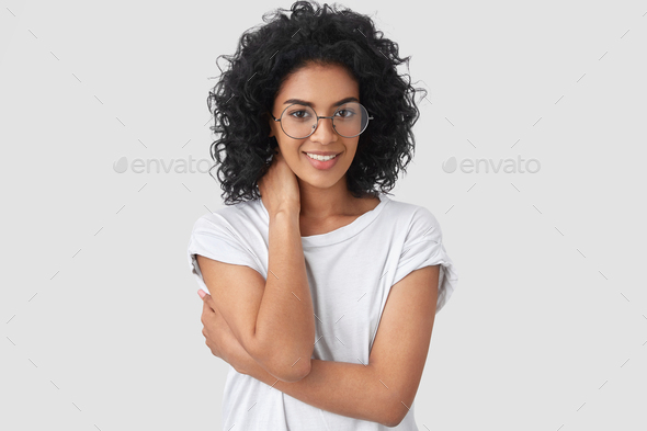 Horizontal shot of shy young mixed race woman with dark skin, curly hair, looks positively, keeps ha