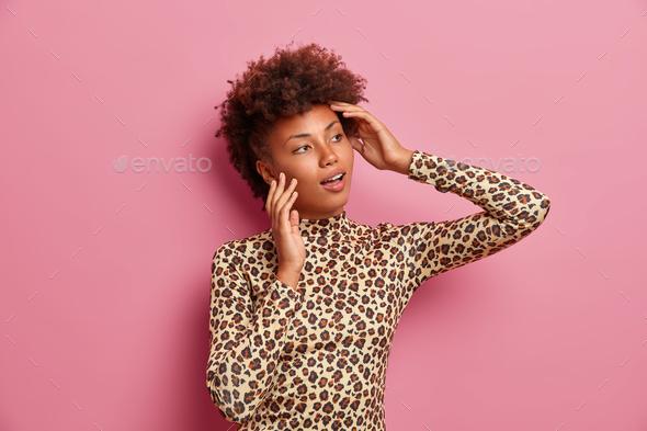 Dark skinned woman with curly hair, looks aside thoughtfully, dressed in stylish leopard outfit, thi