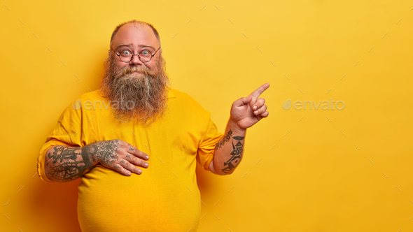 Smiling fat bearded man in eyewear with bald patch and tattooed arms, points on empty space aside, h