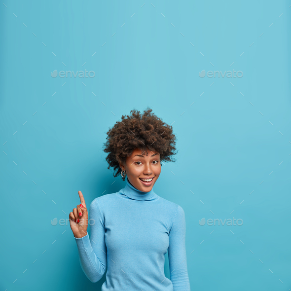 Joyful self confident woman with curly hair points index finger above, shows copy space over head, s