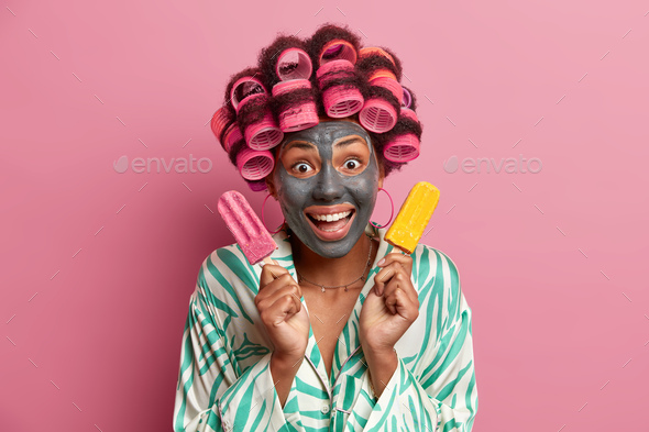 Young cheerful woman applies clay mask for healthy skin and reducing fine lines, poses with deliciou