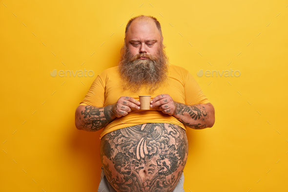 Serious bearded man with big tummy, tattooed arms and belly, holds very small carton cup of coffee c