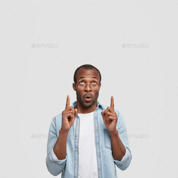 Vertical shot of astonished African American male points upwards with both index fingers, notices so