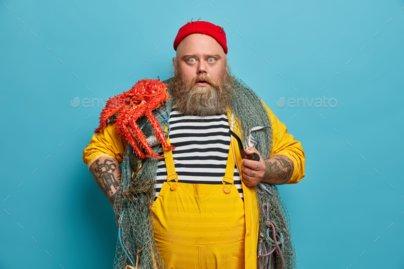 Stupefied bearded man stares bugged eyes at camera holds pipe dressed in yellow overalls red hat car