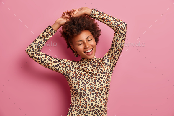 Happy dark skinned woman shakes body, raises hands and dances carefree, wears leopard jumper, closes