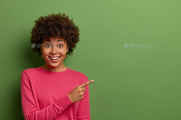 Excited good looking woman with Afro hair points finger right, sees good offer, suggests click link