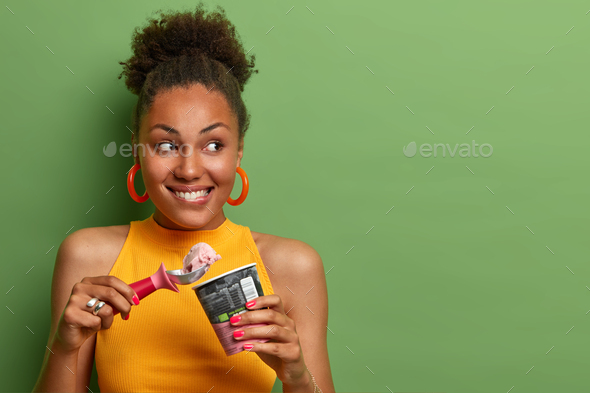 Pleased millennial girl with curly hair, bites lips and eats delicious ice cream with appetite, enjo
