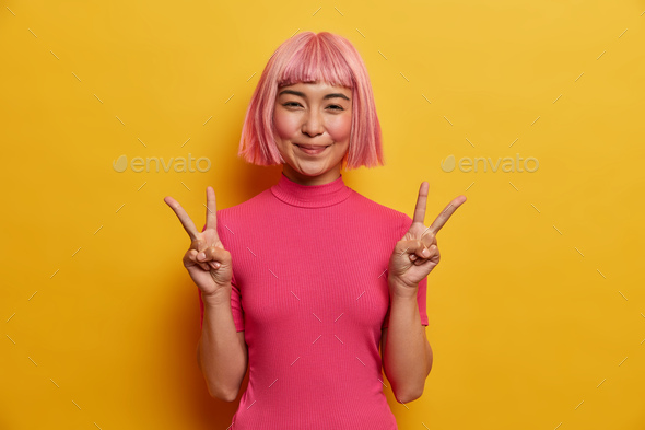 Portrait of happy Asian girl makes peace sign, does victory sign, believes in winning, rejoices succ