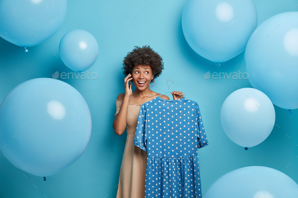 Happy beautiful woman holds blue polka dot dress on hanger, calls someone and uses her phone, prepar