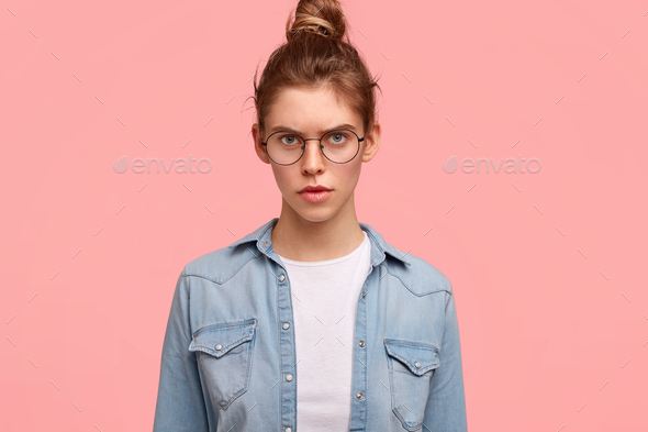 Angry displeased female has sullen expression, dressed casually, wears round spectacles, discontent
