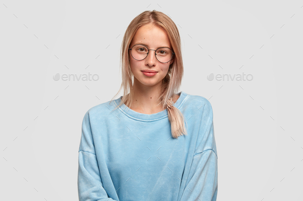 Portrait of beautiful female pupil in oversized sweater, wears spectacles, has pleased expression, h