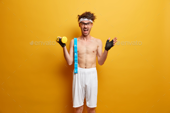 Emotional sportsman lifts heavy dumbbell, shouts emotionally, carries measuring tape, wears sport gl