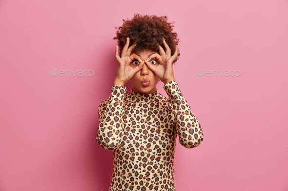 Surprised dark skinned woman in leopard jumper makes goggles with fingers, folds lips and foolishes