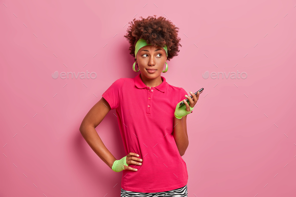 Sporty dark skinned curly haired woman keeps hand on waist, holds mobile phone in hand, uses special