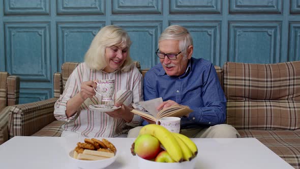 Smiling Senior Couple Grandfather Grandmother Resting on Sofa Drinking Coffee Reading Book at Home alt