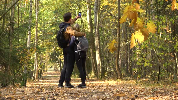 A Hiking Couple Walks Through a Forest on a Sunny Day, Stops To Take a Selfie with a Smartphone alt
