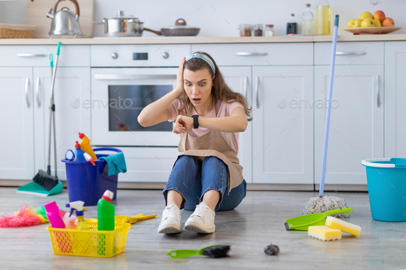 Full length of terrified professional maid sitting on floor at kitchen with cleaning supplies
