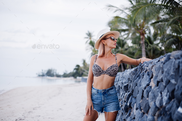 woman in leopard top bikini, jean shorts and classic white hat on tropical exotic beach