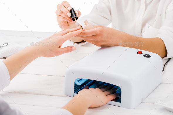 Partial view of manicurist applying nail polish while customer using nail lamp