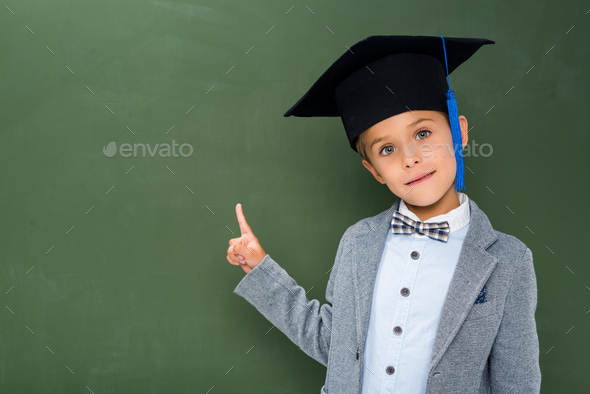 schoolboy in graduation hat pointing at copy space next to chalkboard