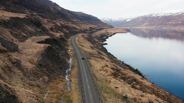 Drone Tracking Car Driving By Fjord And Towards Mountains alt