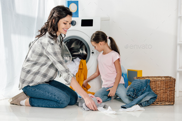 daughter in pink t-shirt and mother in grey shirt gathering clothes on floor and putting in washer
