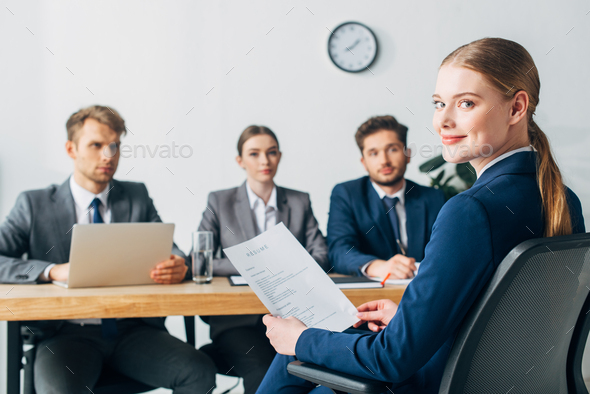 Selective focus of employee looking at camera while holding resume near recruiters