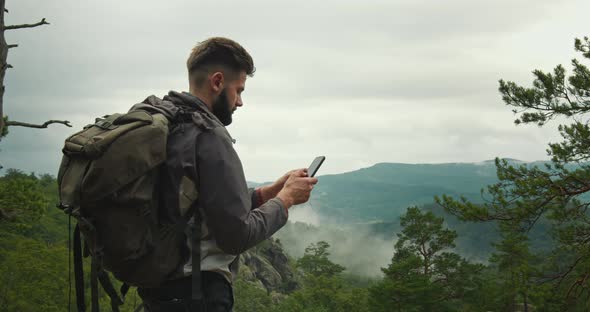 A Man is Standing on Top of a Mountain and Typing a Message on His Smartphone alt