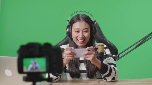 Asian Woman With Headphone Playing Mobile Phone Game While Shooting Video On The Green Screen alt