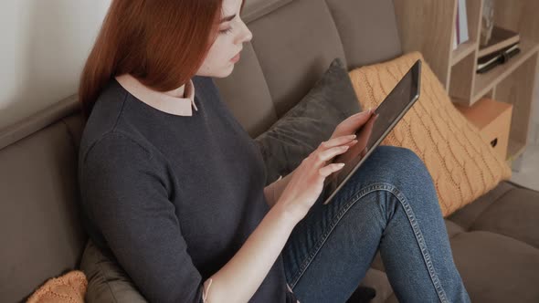Happy young red hair female relaxing at home, playing game on modern tech device. Holding digital ta