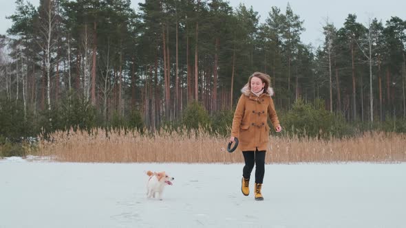 A girl in a coat and yellow boots runs in winter on the ice of the lake with a dog in clothes alt