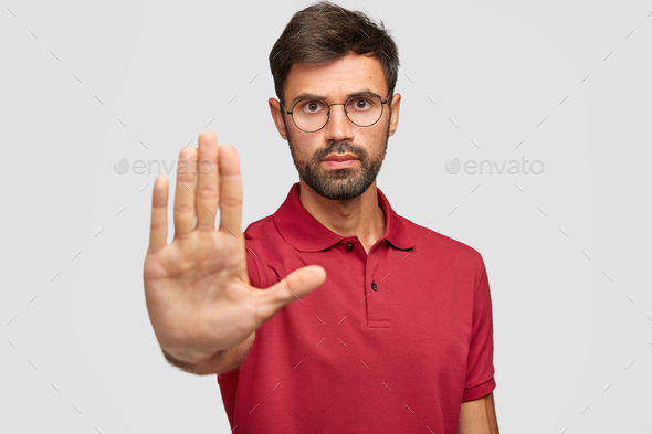 Concerned serious bearded man in round eyewear pulls palm towards camera, stops or warns you from ma