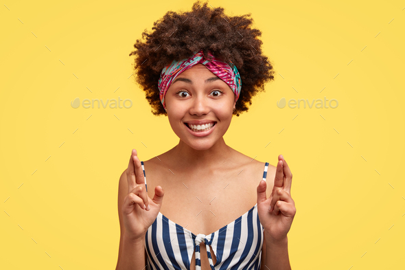 Studio shot of black woman with cheerful expression, crosses fingers, wishes to be lucky for future