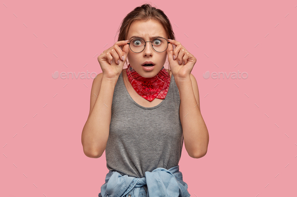 Emotive European woman sees something unbelievable, keeps hands on rim of spectacles, wears red band