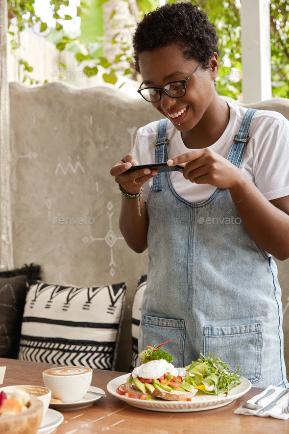 Cheerful black woman makes photo of exotic food in cafeteria, drinks latte, shares in social network