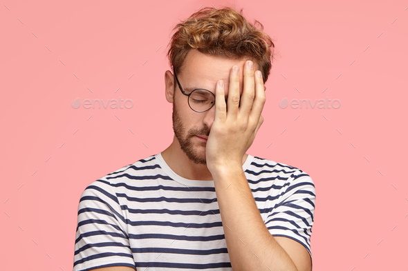 Tired overworked curly ordinary man keeps hand on face, closes eyes, feels sleepy after working all