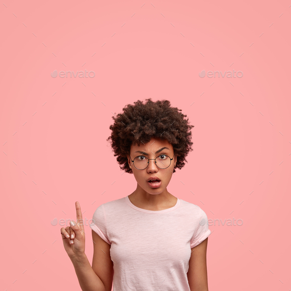 Indignant dark skinned curly lady points with index finger upwards, attracts your attention to blank