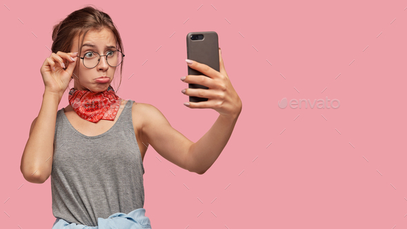 Upset gloomy girlfriend purses lips, makes selfie portrait with modern cell phone, keeps hand on rim