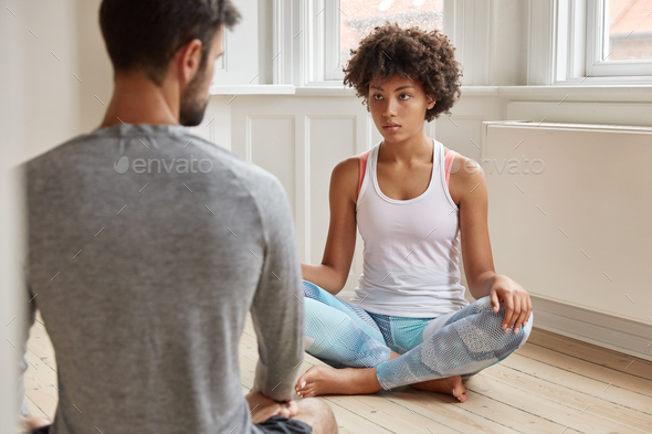 Back view of unshaven man has talk with dark skinned woman, sit on floor together in lotus pose in e