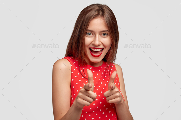 Studio shot of glad young female with positive expression points at camera with both index fingers,