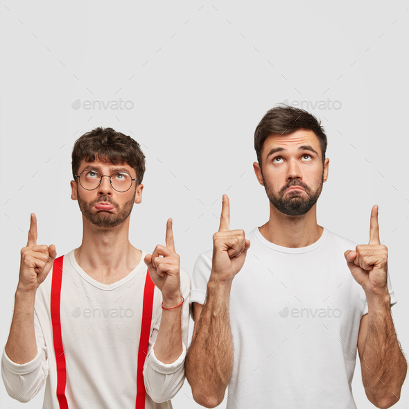 Photo of sullen men with dark hair, dressed in white clothes, points upwards, have displeased facial