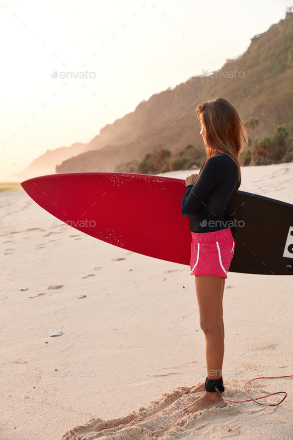 Photo of sportswoman has good body shape, wears pink shorts, carries long surfboard, stands on sandy