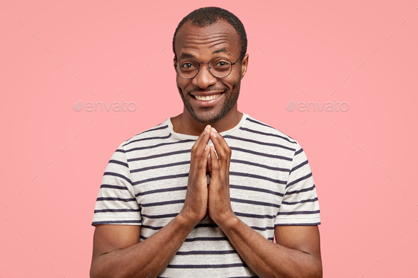 Pleased black man with satisfied expression makes praying gesture, smiles positively at camera, has