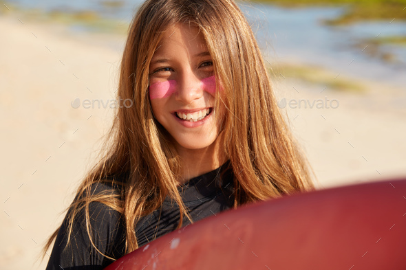 Shot of pleased light haired woman with toothy smile, has surf zinc on face for sun protection, happ