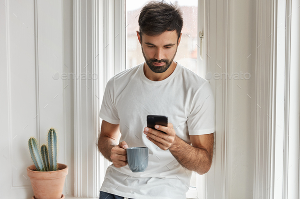 Horizontal shot of handsome bearded man uses modern smart phone device for updating profile in socia