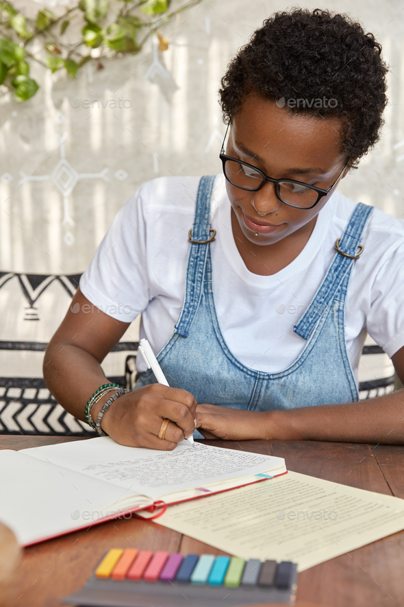 Black woman with boyish haircut, writes in notebook with pen, tries to complete course work, sits at