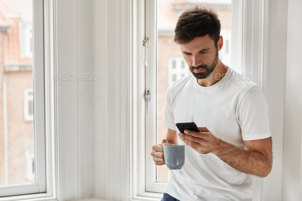 Handsome hipster with beard, types sms message on digital smart phone, dressed in casual t shirt, up