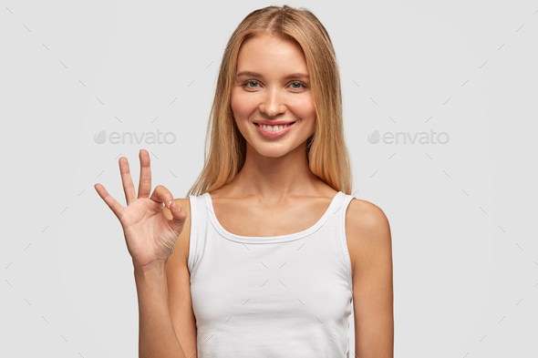 Attractive Caucasian female with long hair, satisfied expression, shows okay sign, feels glad after